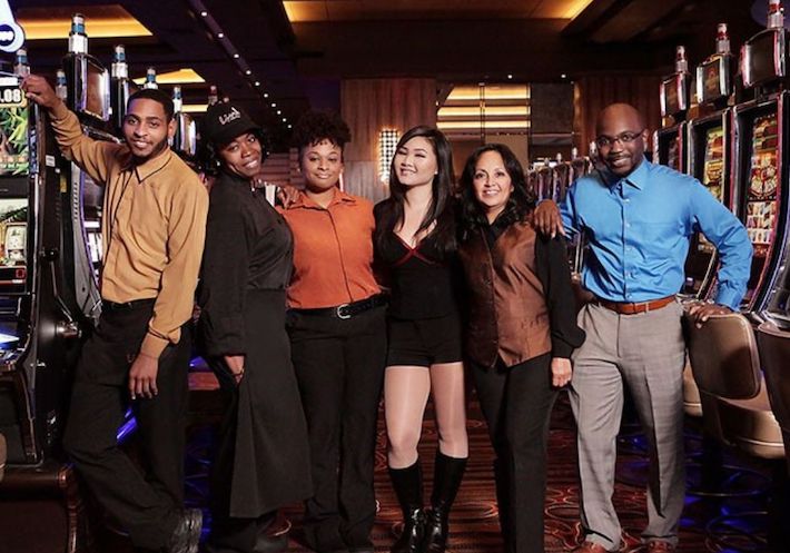 Six diverse casino and hotel workers stand smiling beside rows of slot machines, each wearing dressy and distinctive outfits.
