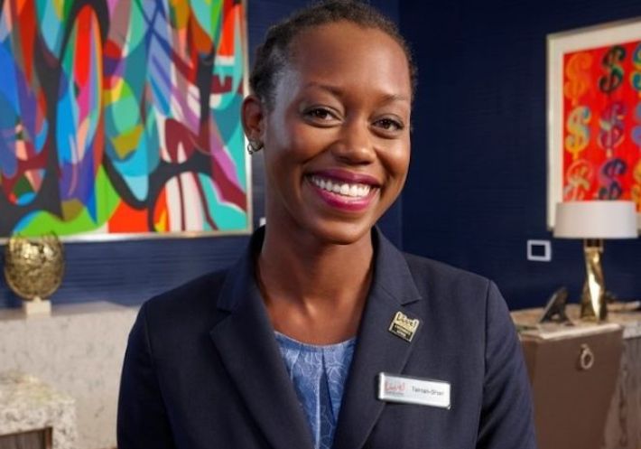 An African-American woman wearing a blazer and Live! pin smiles happily in a blue room with vibrant pictures and a gold vase.