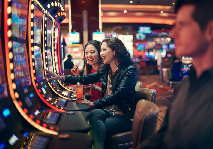 Two women sit smiling at a slot machine, one is pointing at the screen while holding a pink cocktail in her other hand.