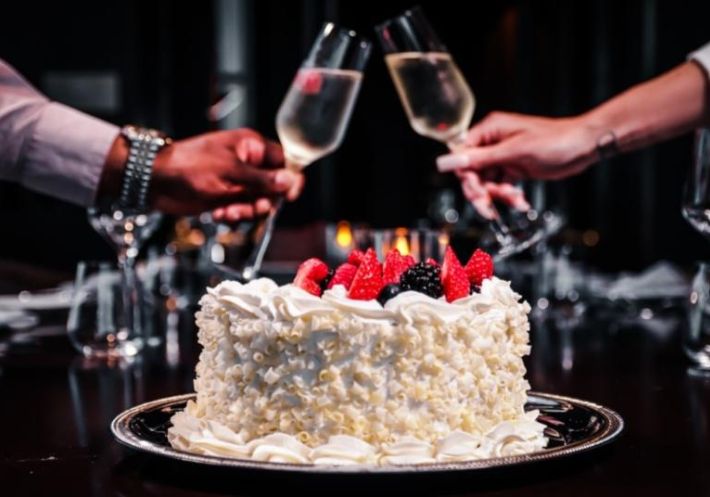 A couple toasts champagne glasses above a delectable white cake decorated with white chocolate curls and topped with berries.